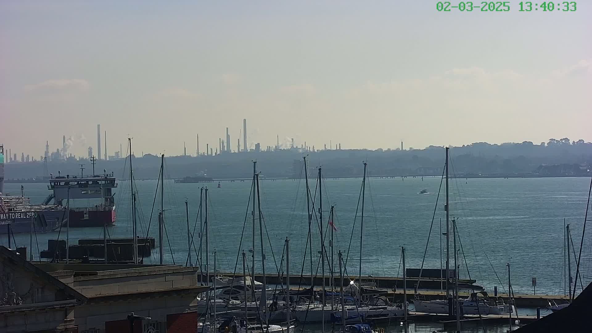 A hazy day reveals a marina filled with sailboats, a ferry, and an industrial plant on the horizon.