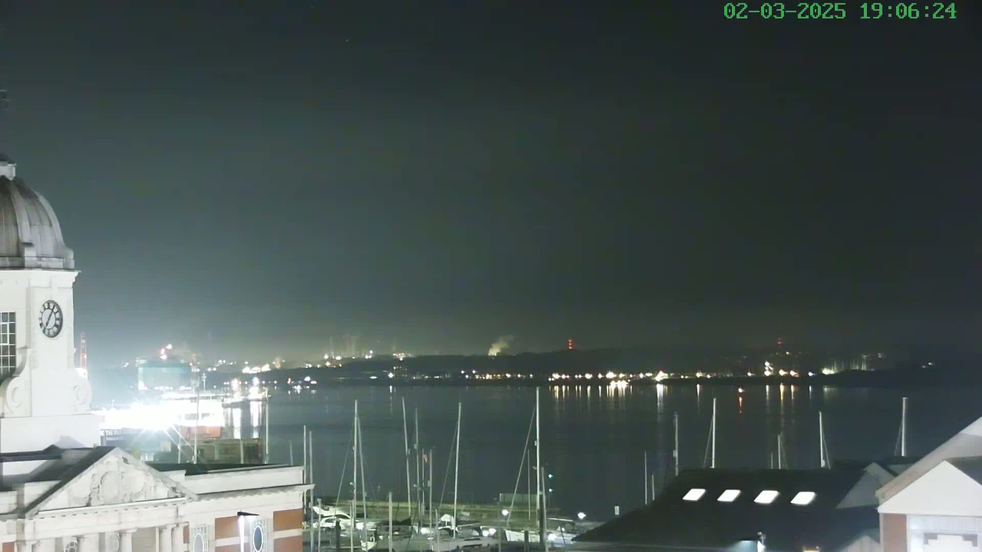 A nighttime view from atop a building shows a calm body of water with numerous sailboats moored, a distant city skyline faintly illuminated, and a portion of a clock tower in the foreground under a dark, clear sky.