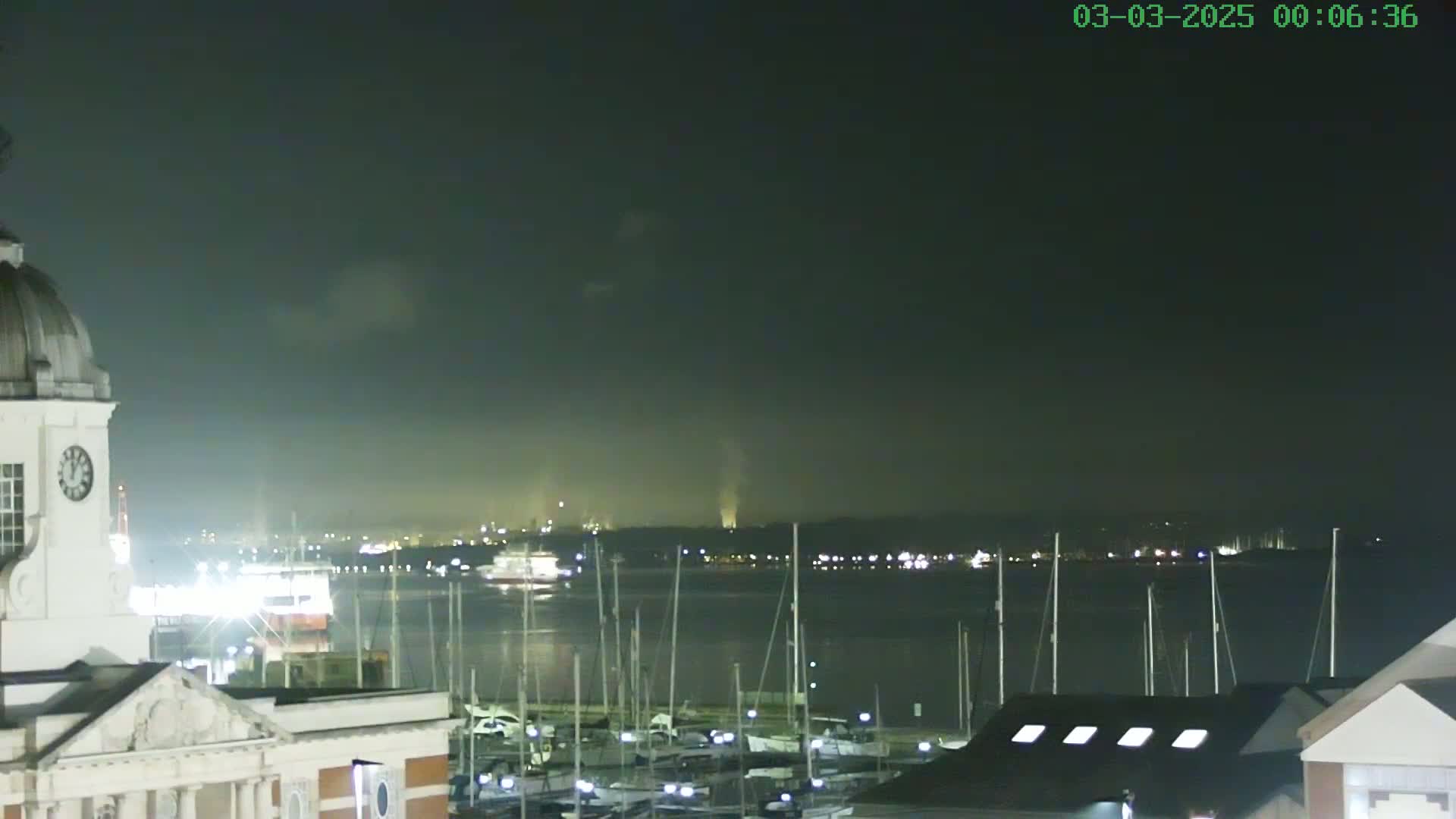A nighttime view of a harbor filled with docked sailboats, a body of water, and a distant shoreline under a dark, cloudy sky.