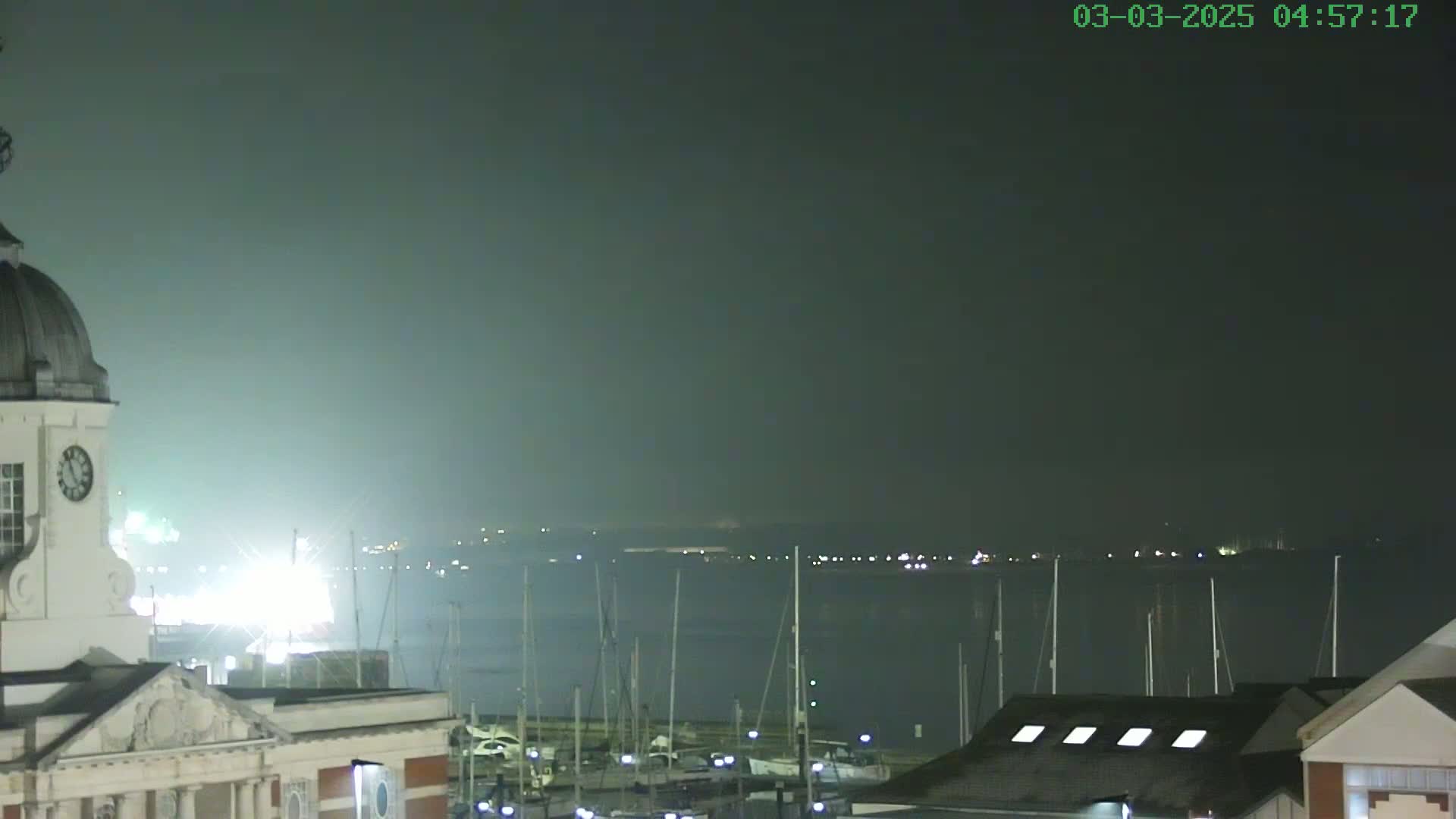 A nighttime view of a harbor filled with docked sailboats, under hazy conditions, with a clock tower visible in the foreground.