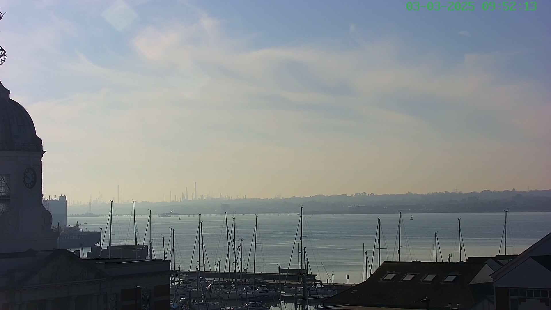 A hazy, sunny day reveals a harbor filled with sailboats, a distant industrial skyline, and a portion of a clock tower.
