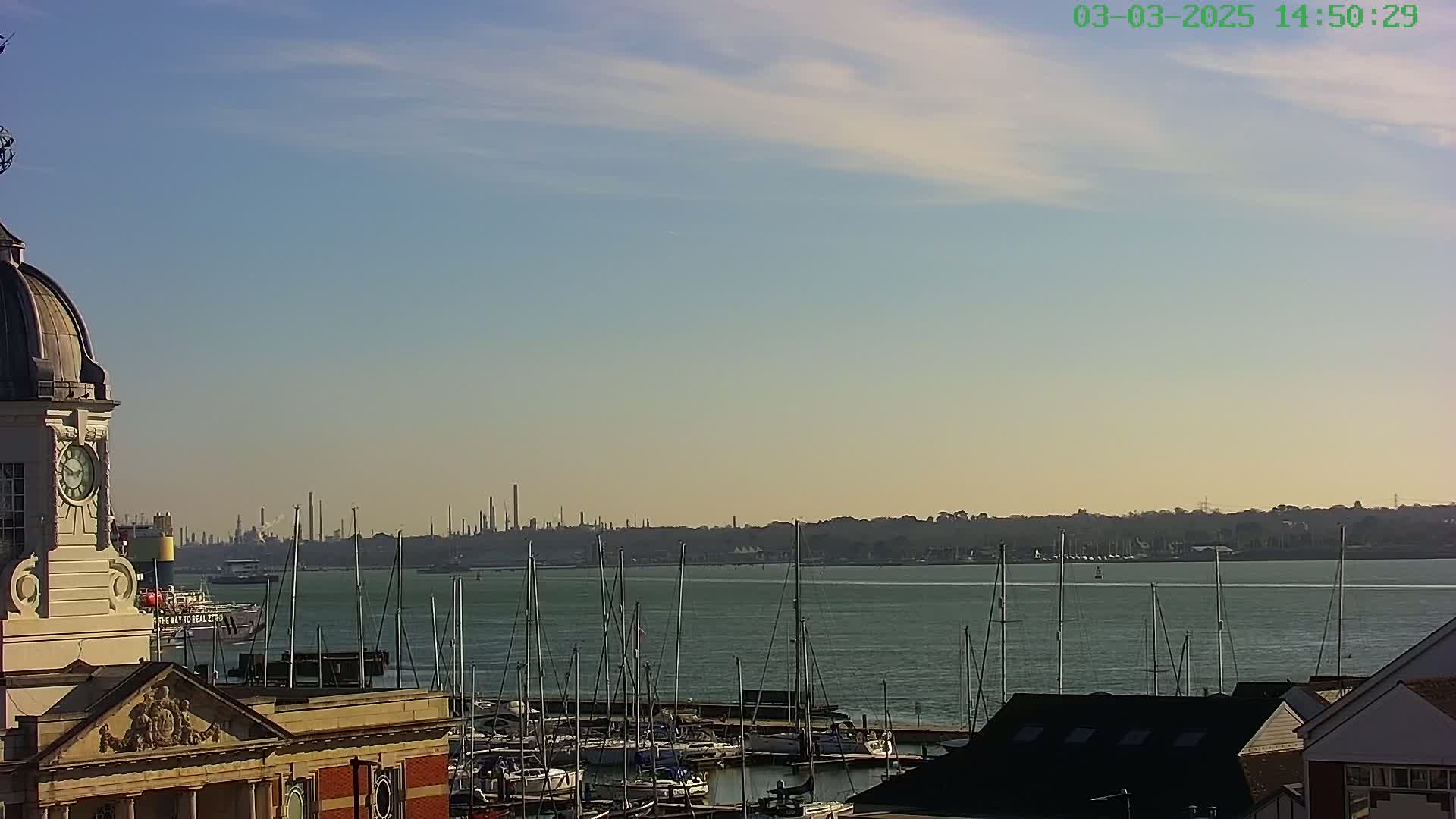 A clock tower overlooks a marina filled with sailboats, with a hazy industrial skyline visible across the calm water under a mostly clear sky.