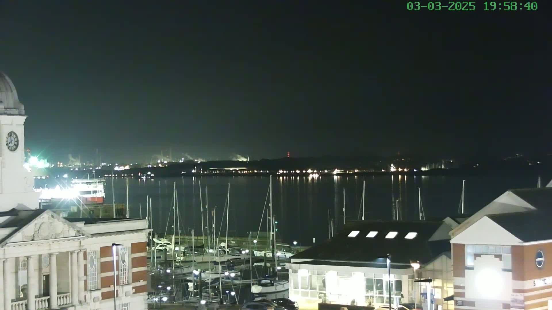A nighttime view of a harbor with many sailboats docked, a large building with a clock tower on the left, and other buildings in the foreground, under a dark, clear sky.