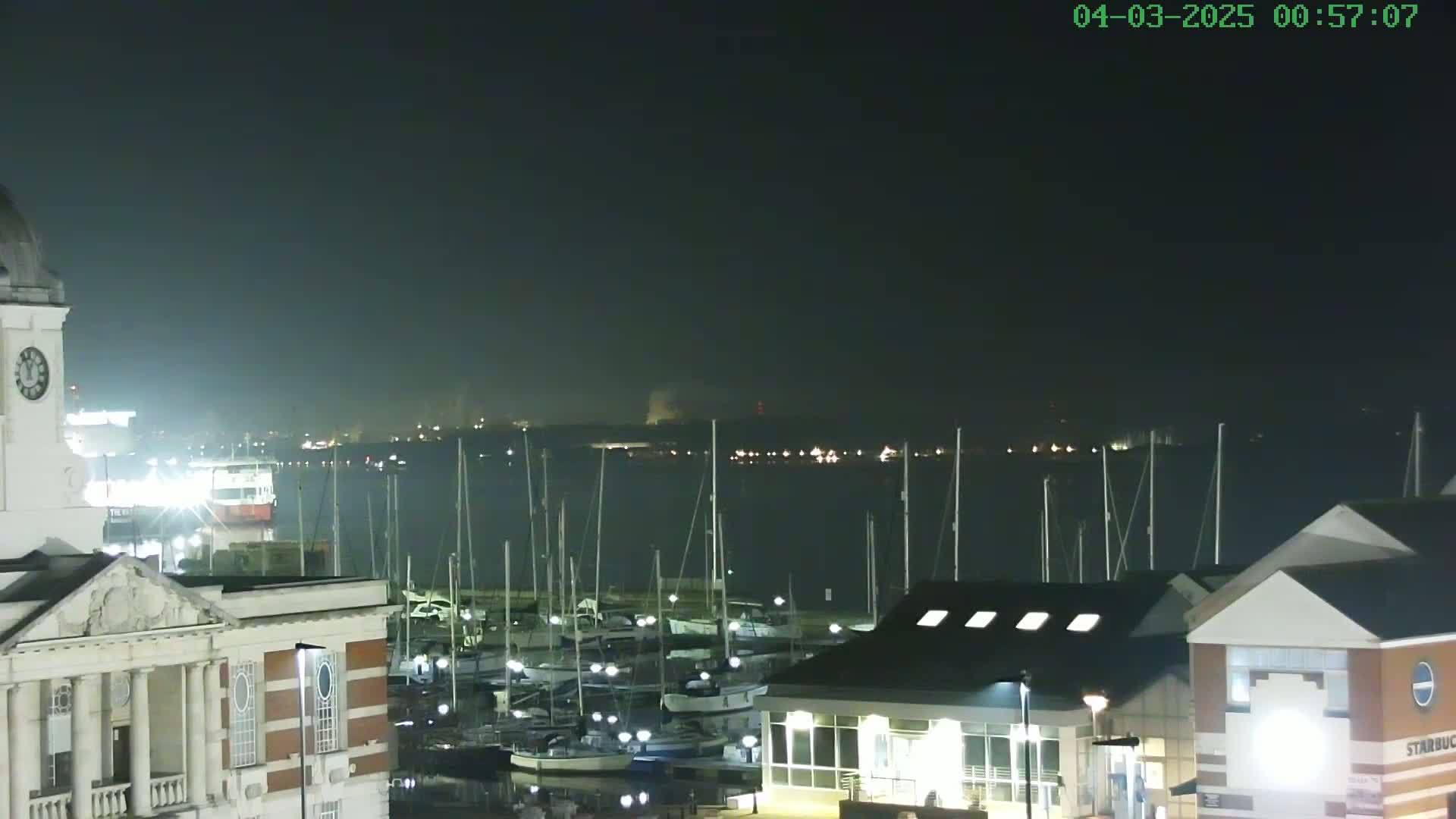 A nighttime view of a harbor filled with docked sailboats, seen from a vantage point overlooking a building with a clock tower.