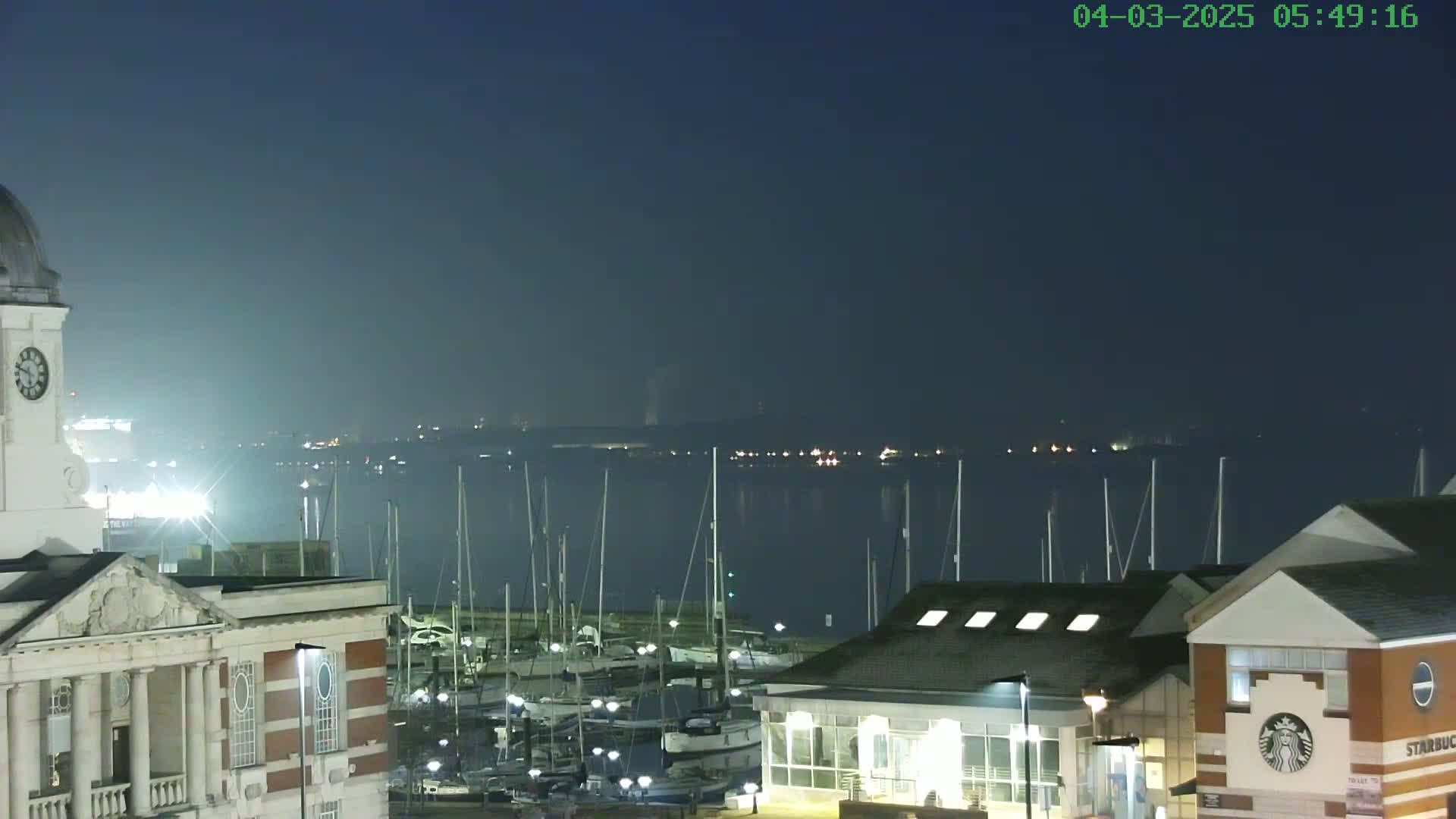 A nighttime view of a harbor filled with sailboats, overlooked by buildings, under a dark, clear sky.