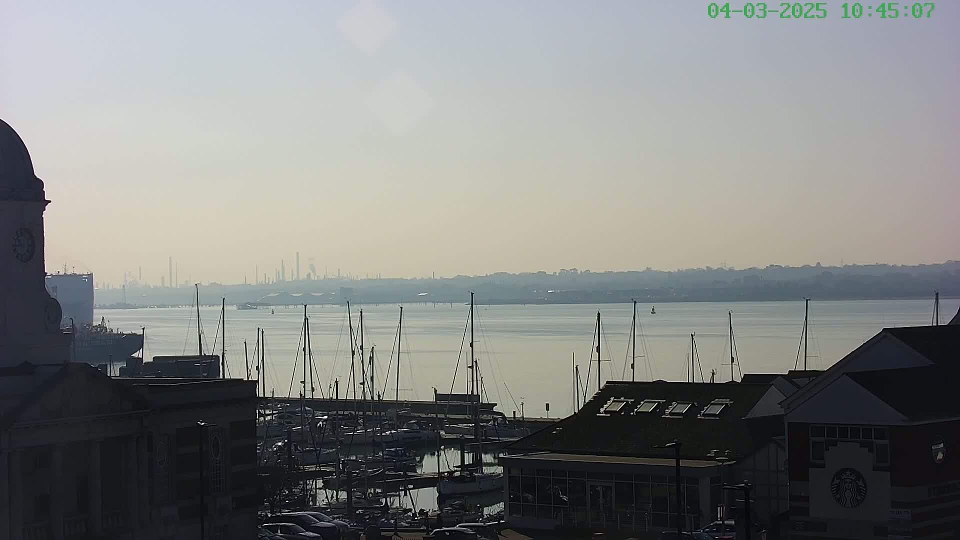 A hazy, sunny day overlooking a marina filled with sailboats, with a distant industrial skyline visible across the calm water.