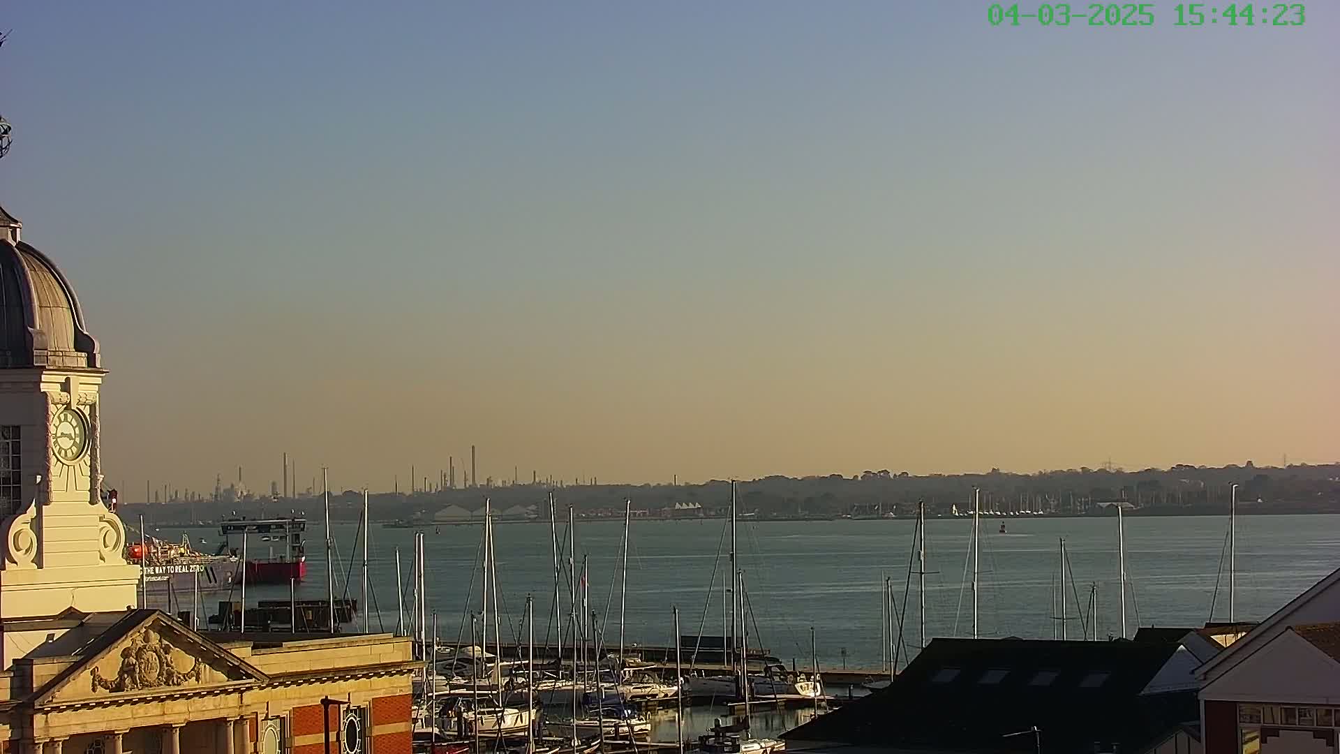 A clock tower overlooks a marina filled with sailboats, with an industrial skyline visible across a calm body of water under a clear, pale sky.