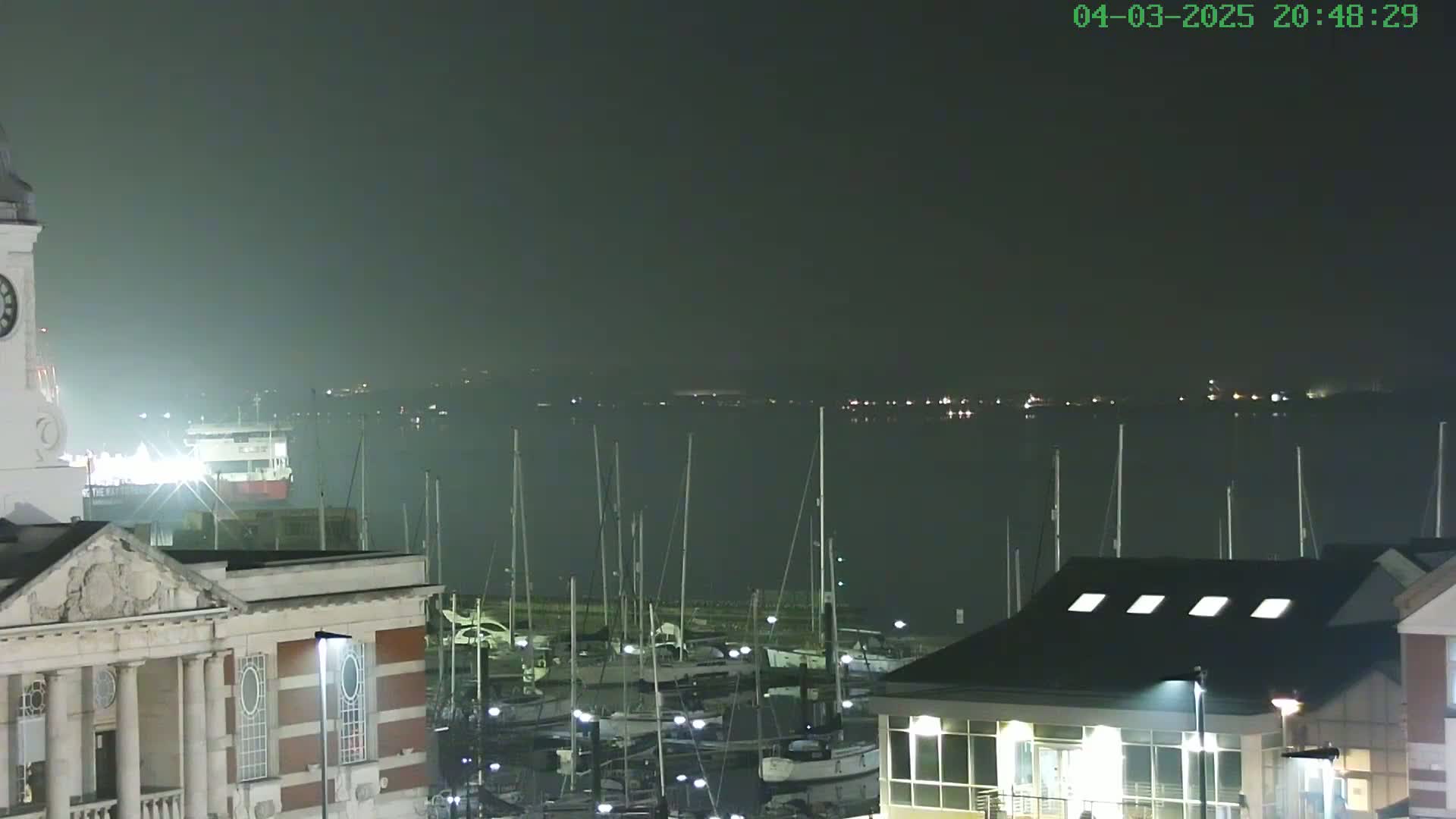 A nighttime view of a harbor filled with docked sailboats, a large building with a clock tower, and a hazy atmosphere over the water.