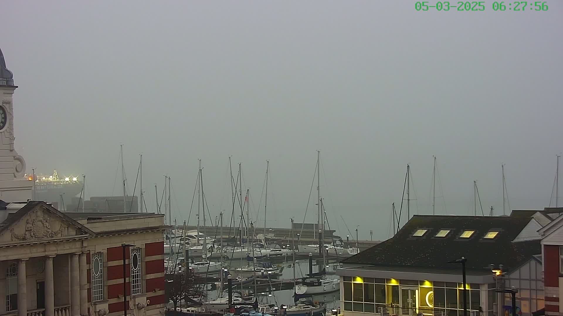 A foggy harbor scene shows numerous sailboats moored at a dock, viewed from a vantage point near a brick building with a clock tower.