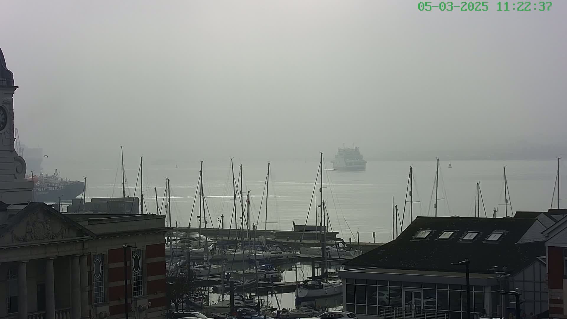 A ferry is visible in the distance on a foggy day over a harbor filled with docked sailboats, near a building with a clock tower.