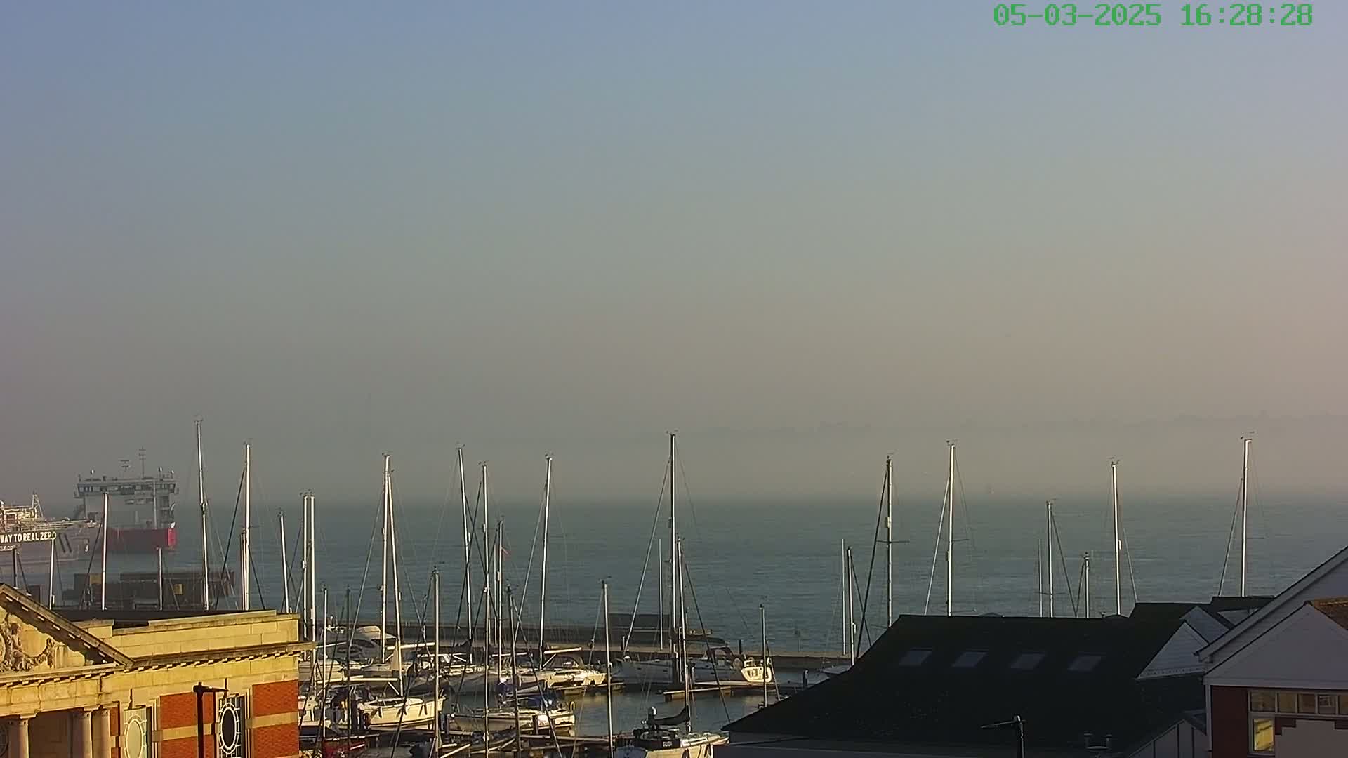 A hazy harbor scene shows numerous sailboats moored at a marina, with a large ship visible in the distance.
