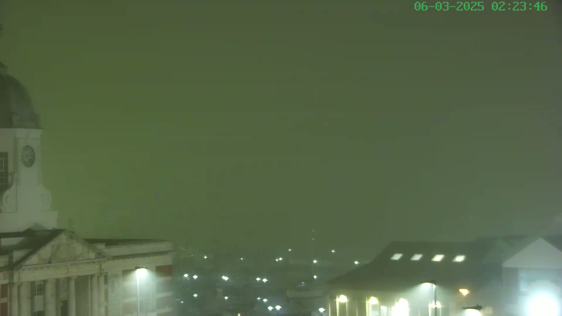 A portion of a city is shrouded in thick fog at night, illuminated by streetlights, with a clock tower visible in the foreground.