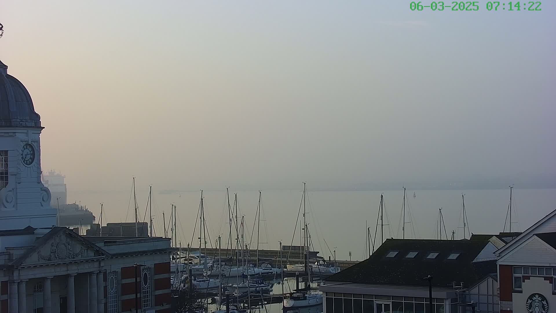A hazy dawn reveals a marina filled with sailboats, overlooked by a building with a clock tower and other structures.