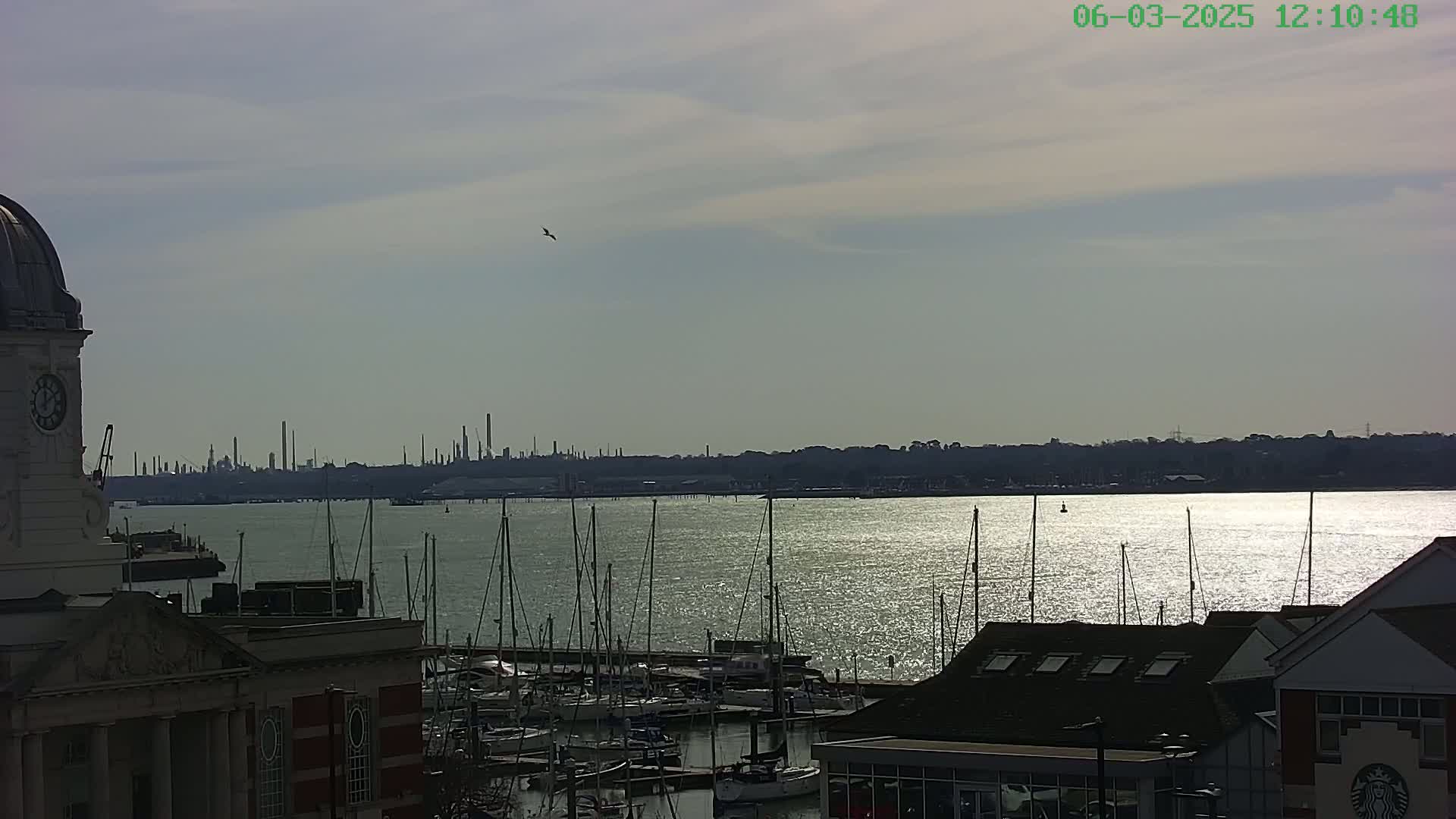 A marina filled with sailboats is situated in front of a body of water reflecting sunlight, with an industrial skyline visible in the distance under a partly cloudy sky.