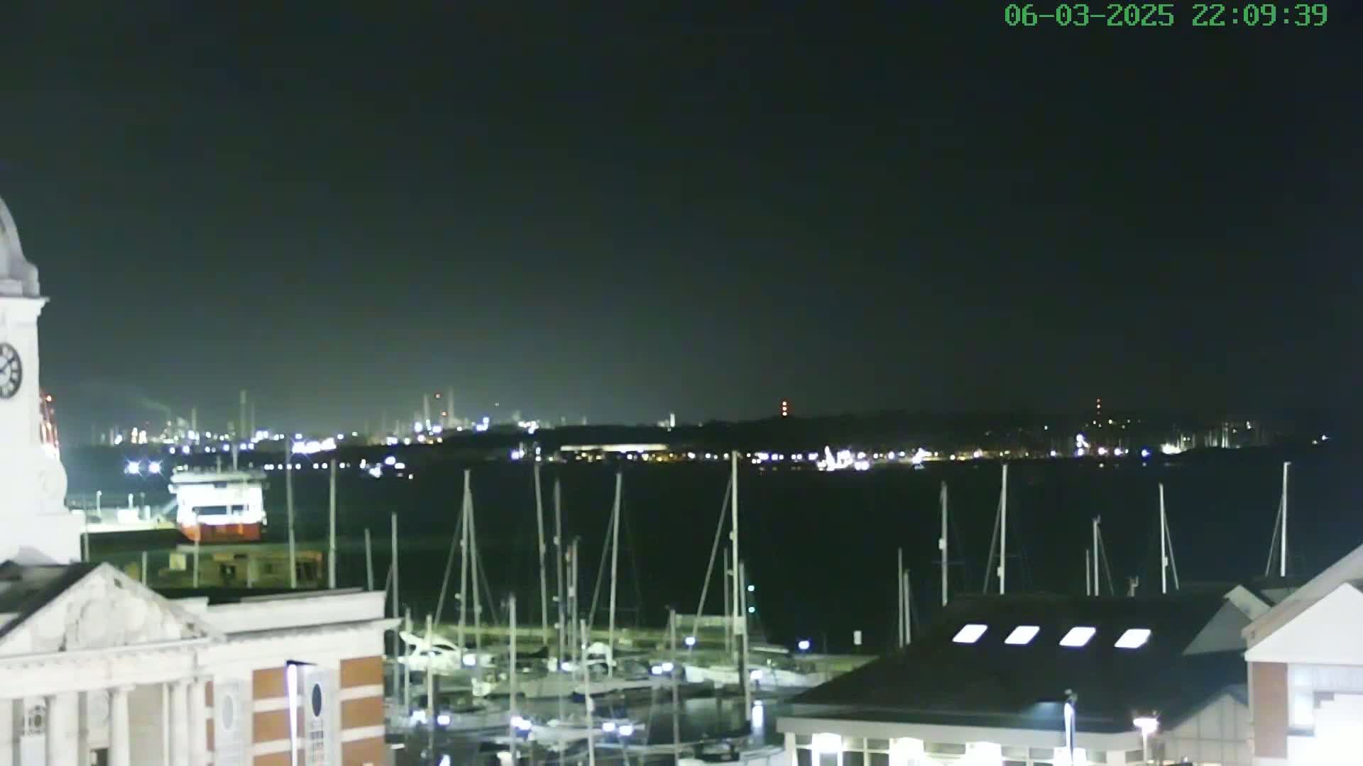 A nighttime view shows a marina with numerous sailboats docked, a portion of a building with a clock tower, and a distant city skyline illuminated under a dark sky.