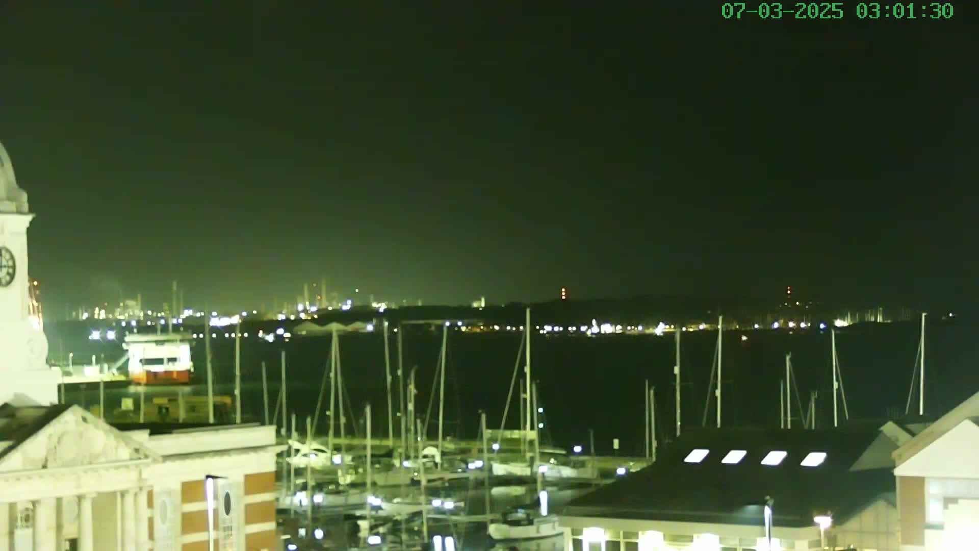 A nighttime view of a harbor filled with docked sailboats, with a city skyline faintly visible in the distance under a dark sky.
