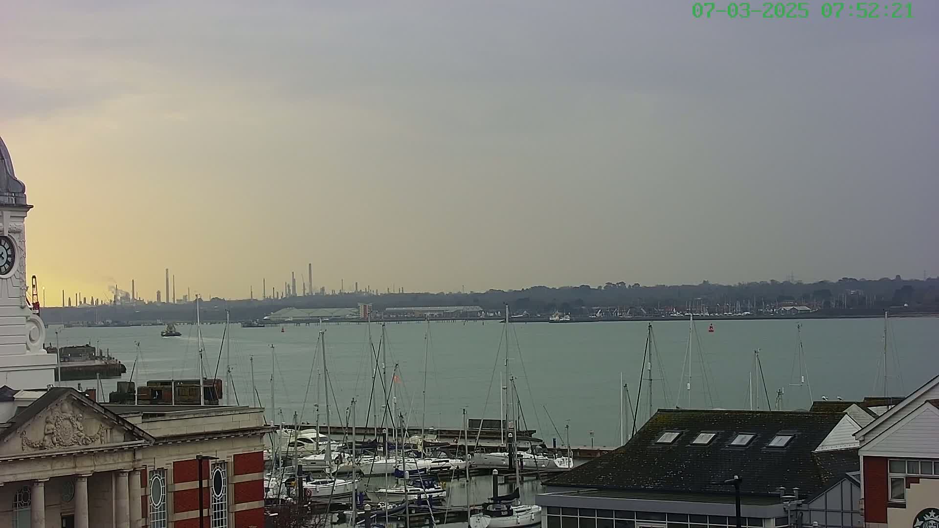 A clock tower overlooks a calm harbor filled with numerous sailboats, with an industrial skyline visible in the distance under an overcast sky.