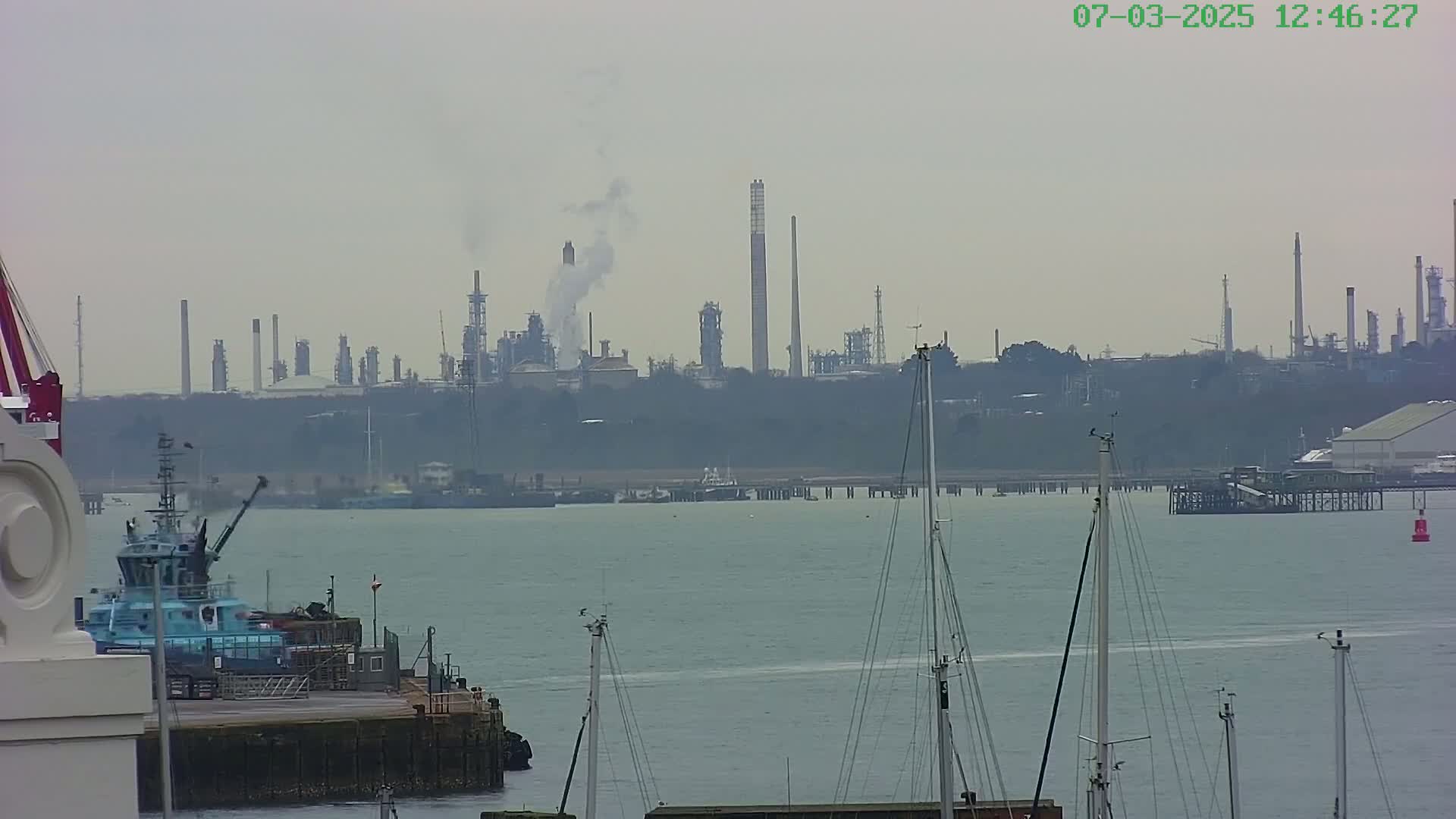 A hazy view of a calm body of water shows docked boats in the foreground and a large industrial complex emitting smoke in the background.
