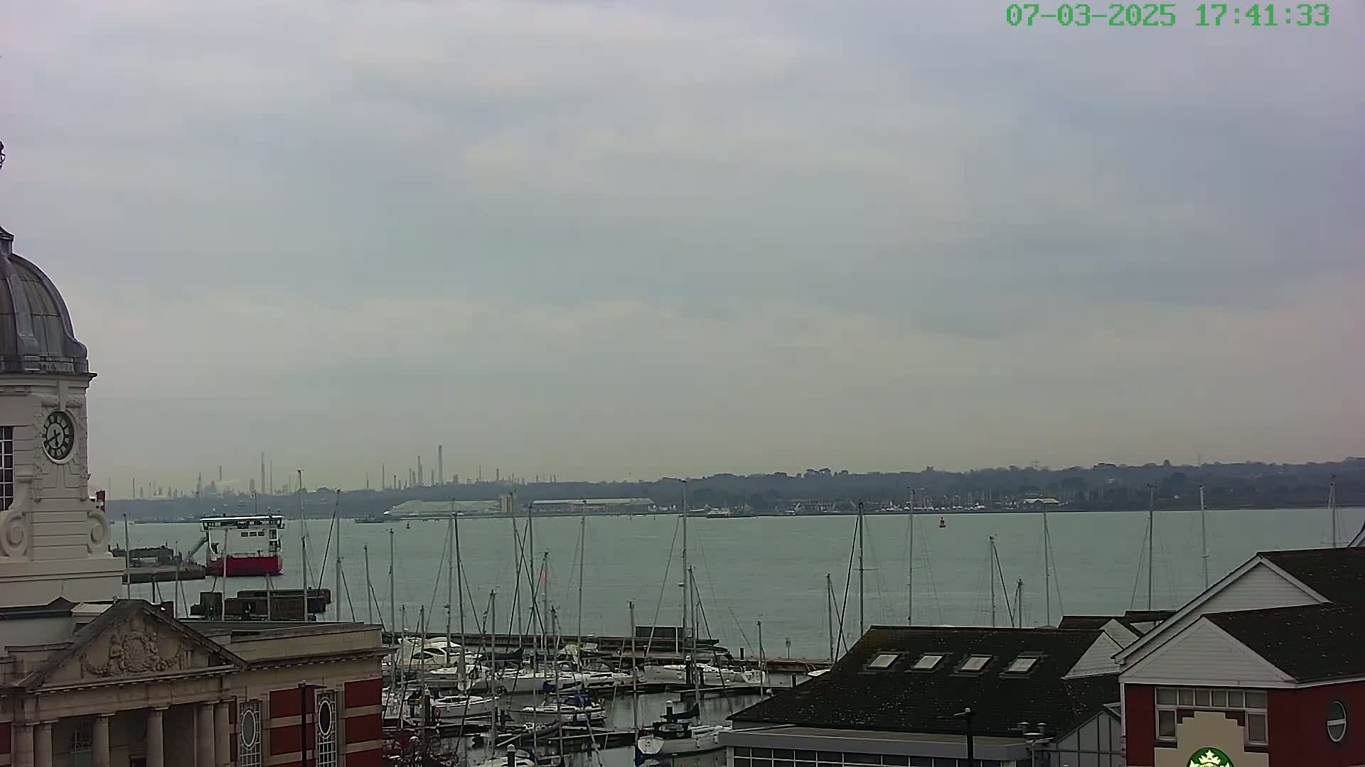 A harbor filled with sailboats is seen from a vantage point overlooking a building with a clock tower, under a cloudy sky, with an industrial skyline visible in the distance.