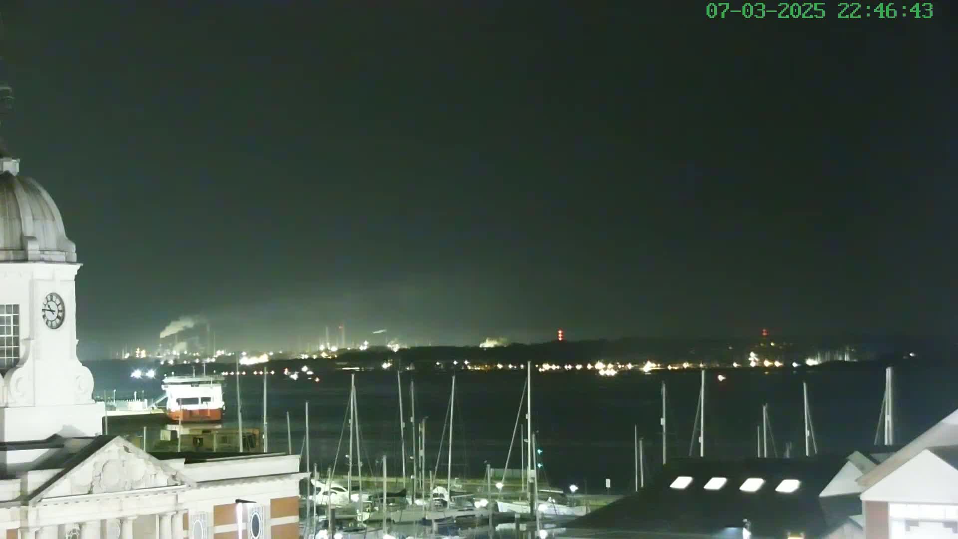 A nighttime view of a harbor filled with docked sailboats, with a distant industrial area illuminated and a clock tower in the foreground under a dark, clear sky.