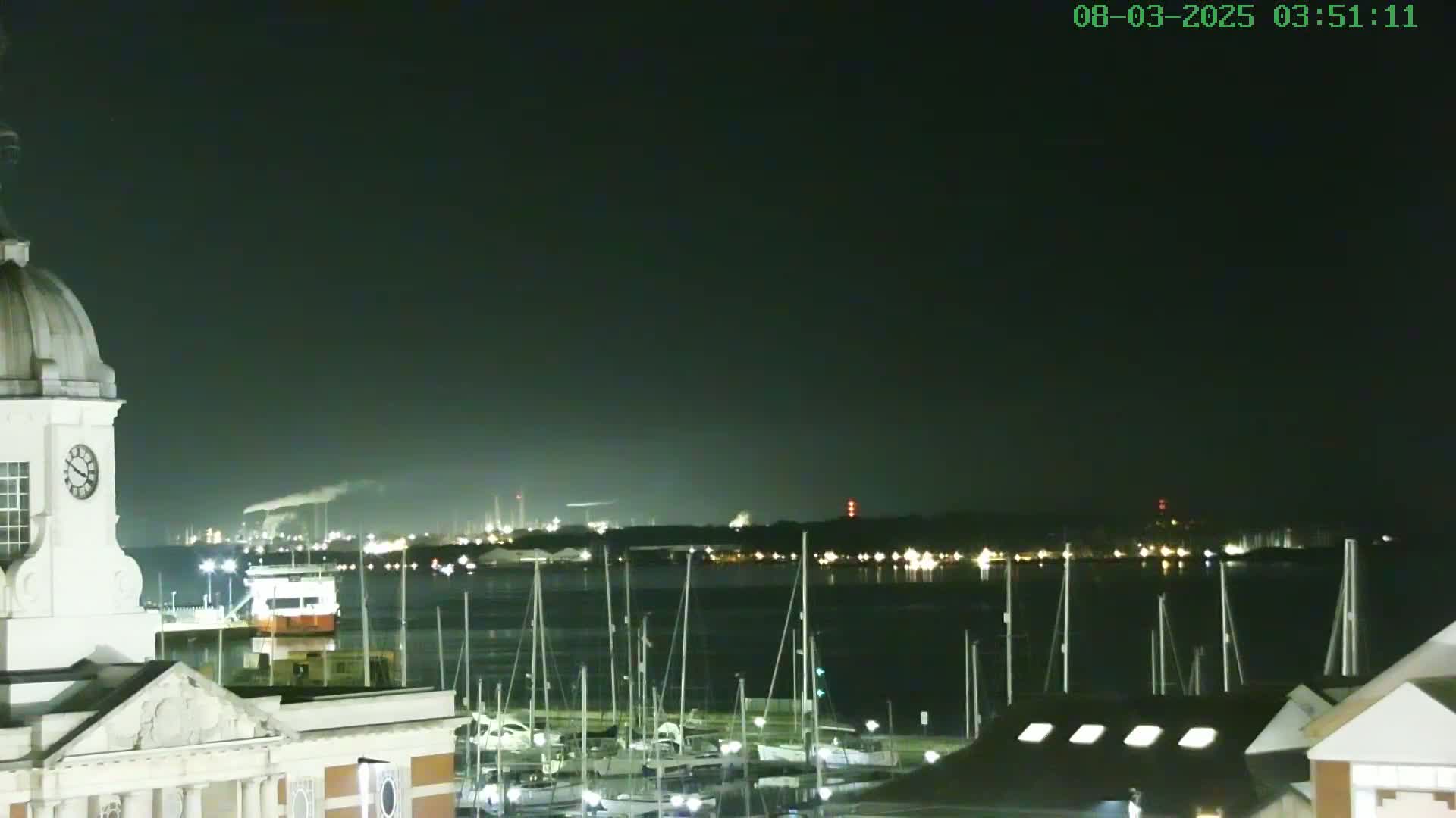 A nighttime view of a harbor filled with docked sailboats, with a clock tower and industrial lights visible in the distance under a dark, clear sky.
