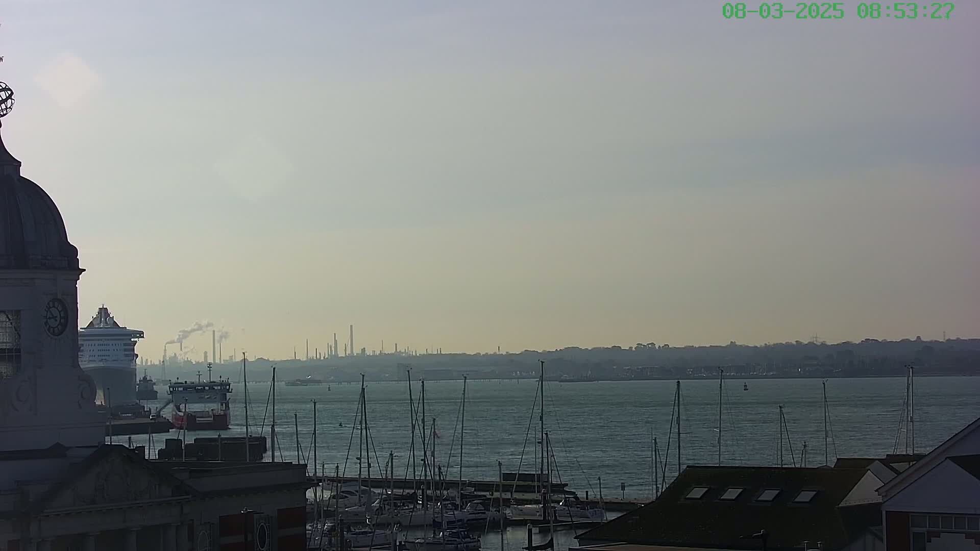 A hazy view of a harbor with numerous sailboats, a cruise ship, and an industrial skyline in the distance, seen from atop a building with a dome.