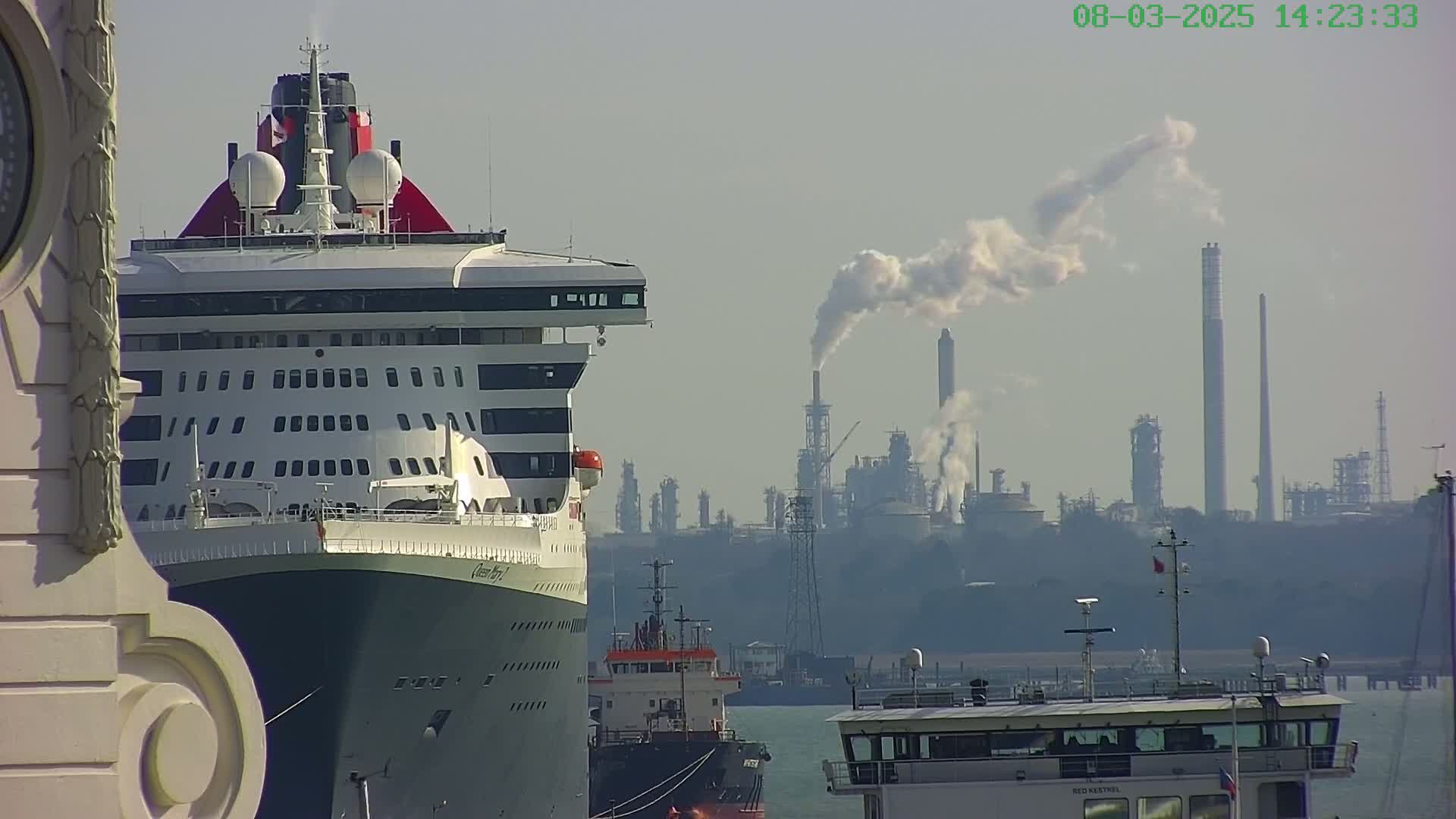 A large cruise ship is docked near an industrial plant emitting smoke on a hazy day.