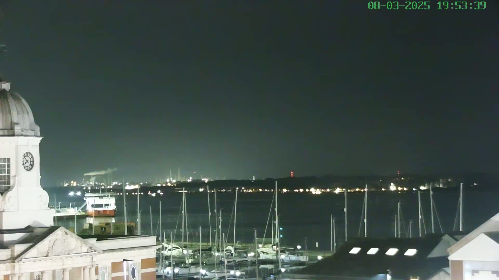 A nighttime view of a harbor with many docked sailboats, a distant industrial skyline faintly visible across the water under a dark sky, and a portion of a white clock tower in the foreground.