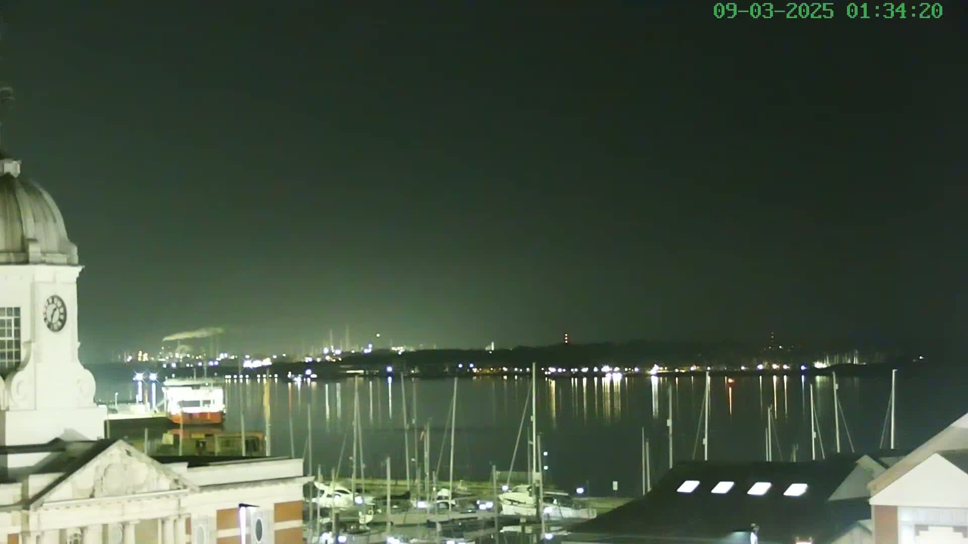 A nighttime view of a harbor filled with docked sailboats, a clock tower, and a cityscape reflected in calm water under a dark sky.