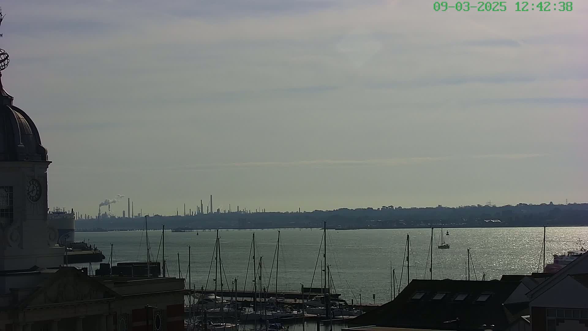 A view from a building shows a calm body of water with numerous sailboats moored in a marina, a distant industrial skyline, and a hazy sky.