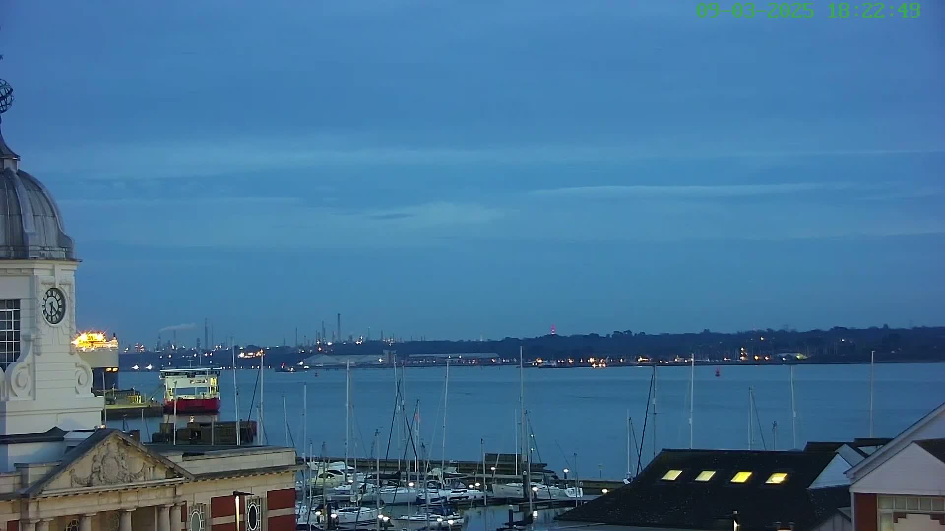 A clock tower overlooks a calm, twilight harbor filled with docked sailboats and a distant industrial skyline.