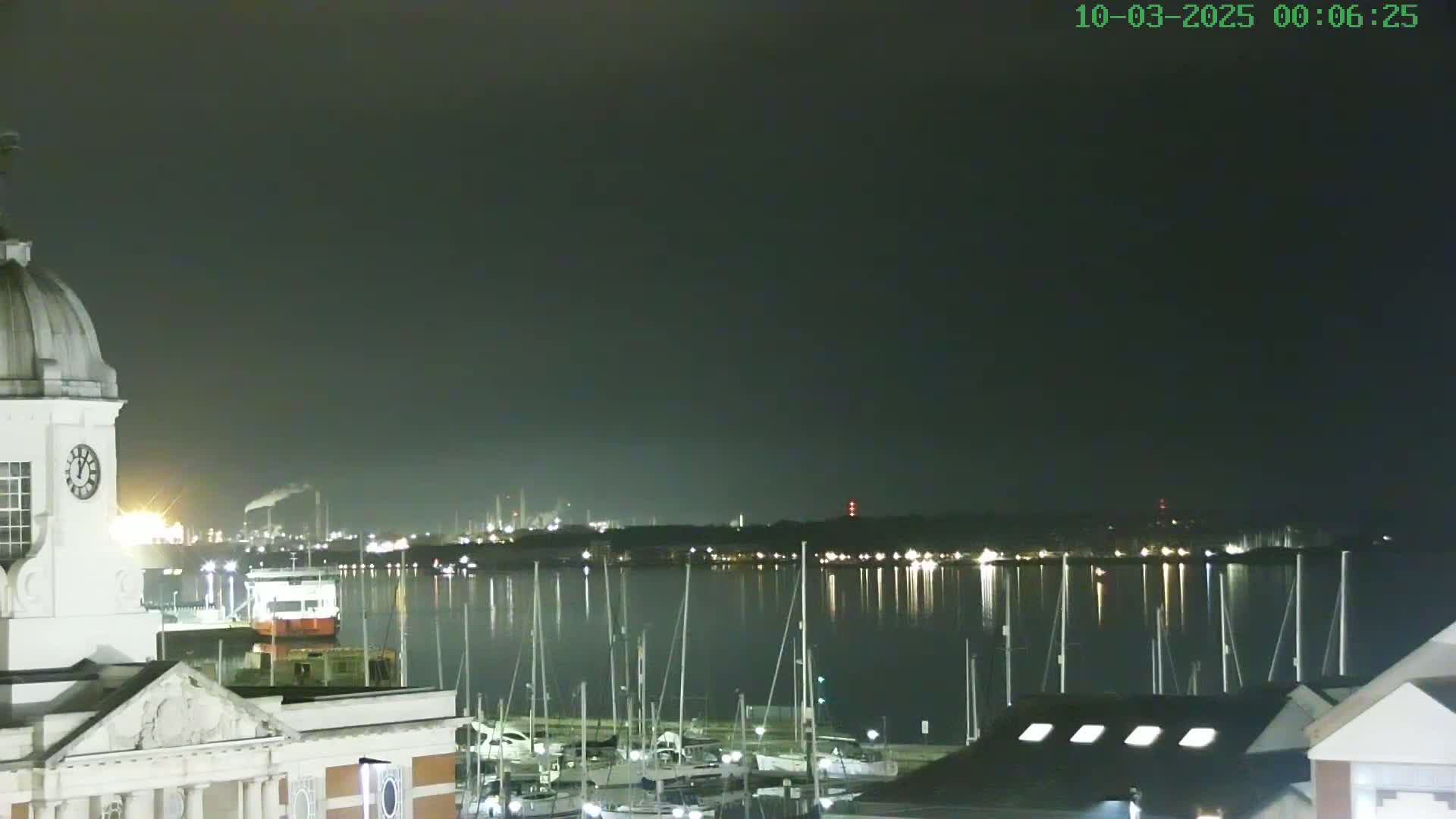 A nighttime view of a harbor filled with docked sailboats, with a portion of a building and a distant industrial area illuminated under a dark sky.