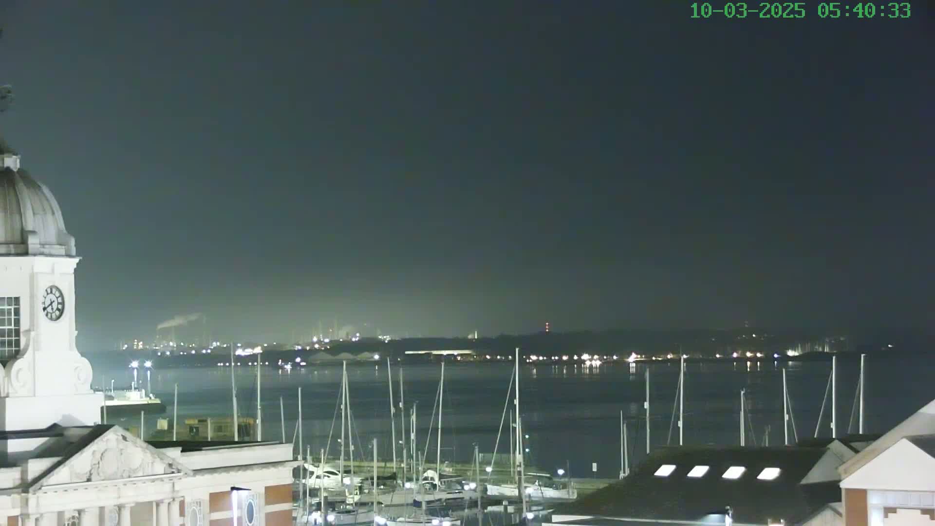 A nighttime view from atop a building shows a calm body of water with numerous sailboats docked, a distant cityscape with faint lights, and a dark, clear sky.