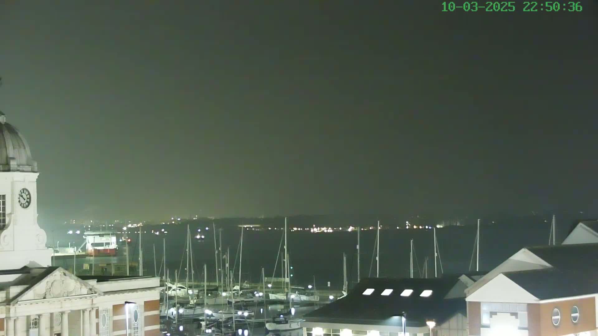 A nighttime view of a harbor filled with docked sailboats, overlooked by a clock tower and modern buildings under a dark, hazy sky.