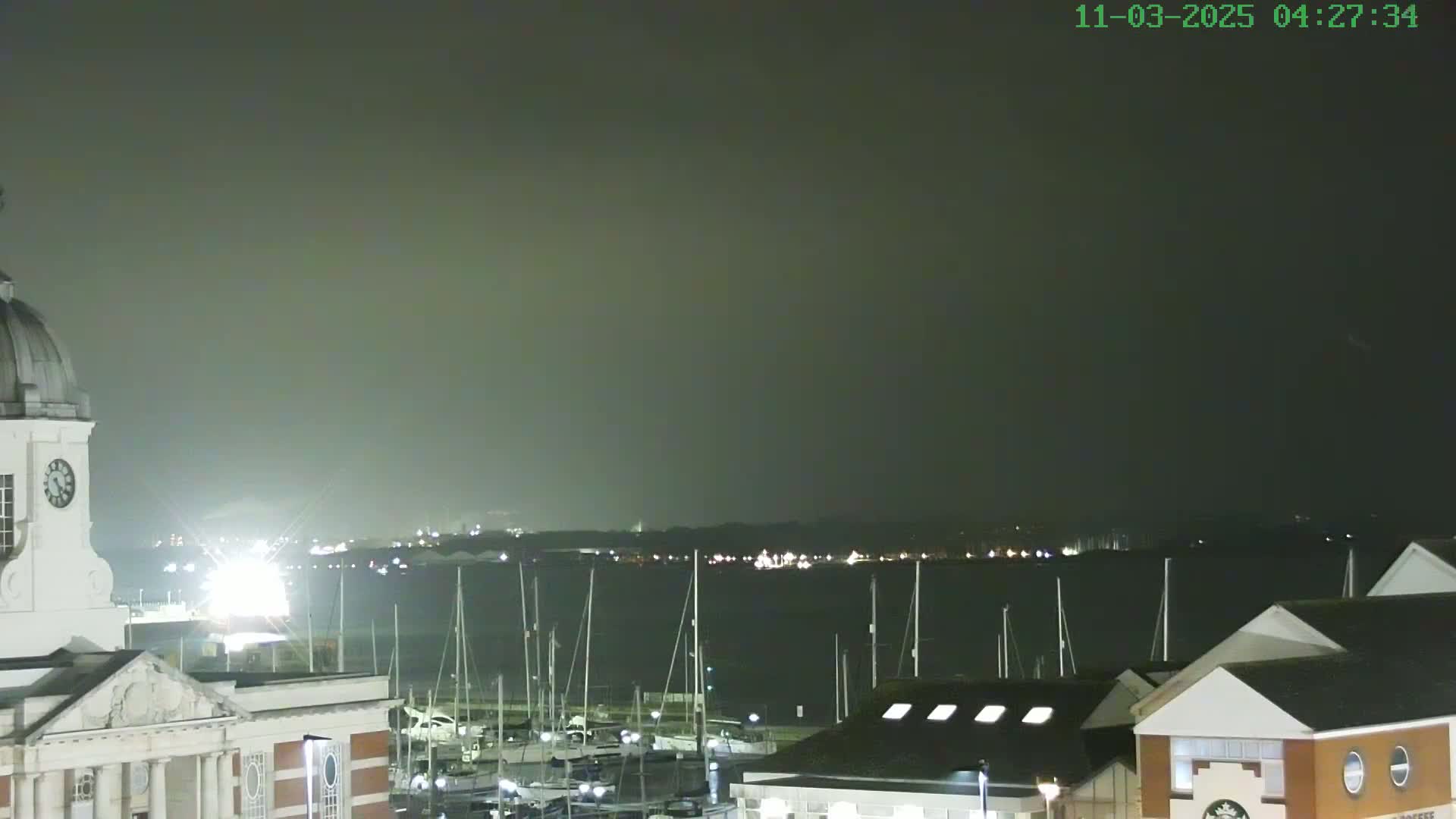 A nighttime view of a harbor filled with docked sailboats, under a hazy, dark sky, with a clock tower and buildings in the foreground.