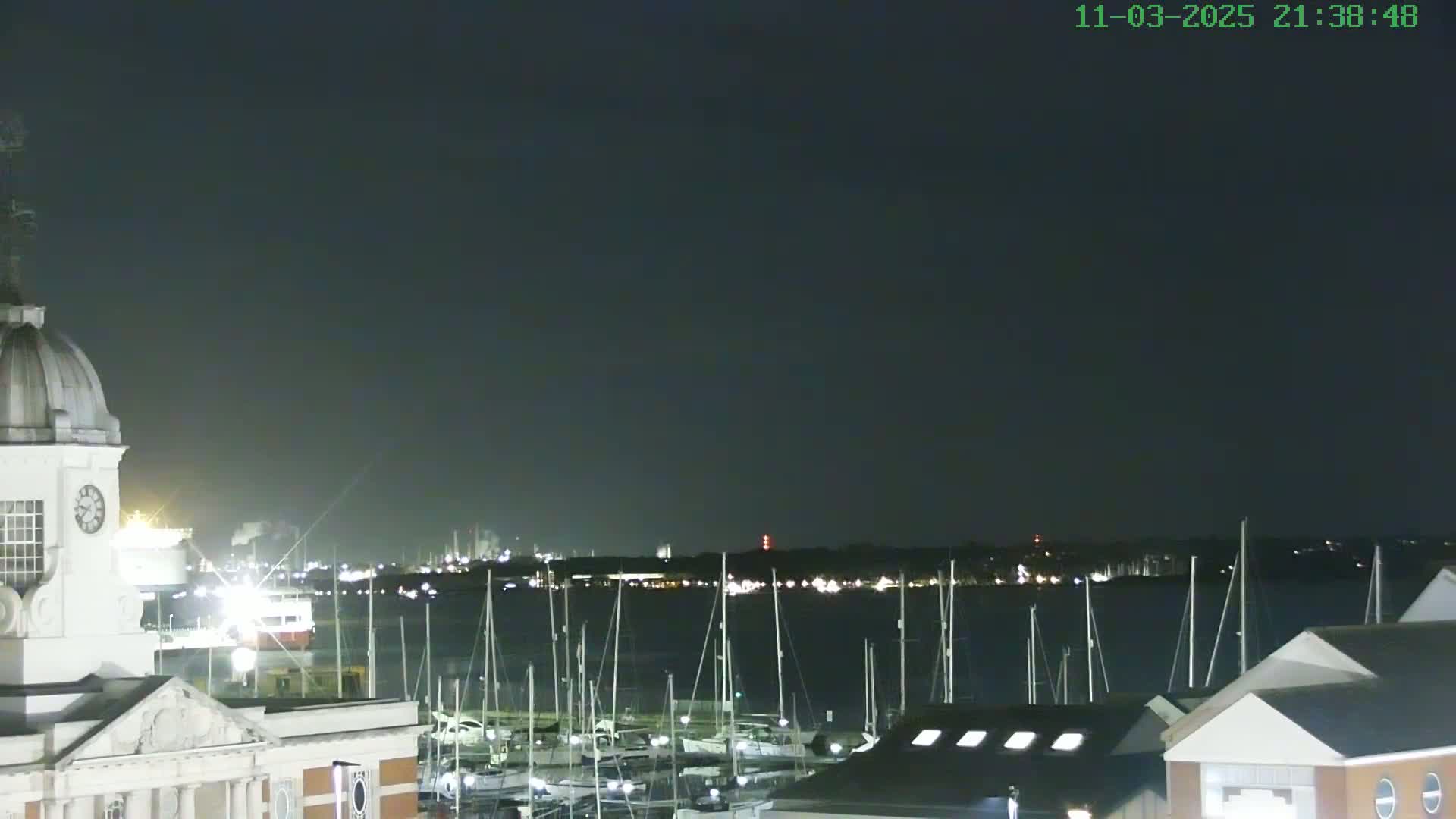 At night, a harbor filled with sailboats is seen from atop a building with a clock tower under a dark sky.