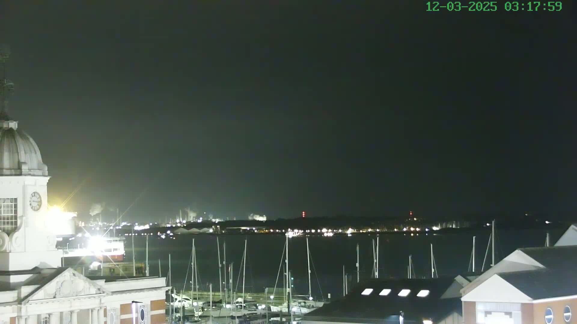 A nighttime view of a harbor filled with sailboats, a distant industrial skyline, and a clock tower atop a building in the foreground under a dark, clear sky.