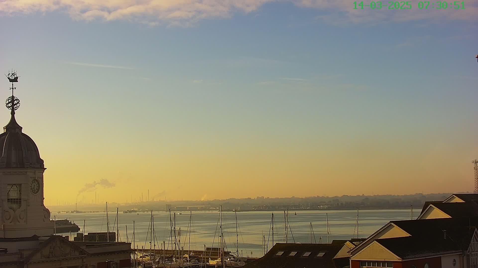 A hazy sunrise illuminates a harbor filled with sailboats, viewed from a vantage point overlooking a clock tower and several buildings.