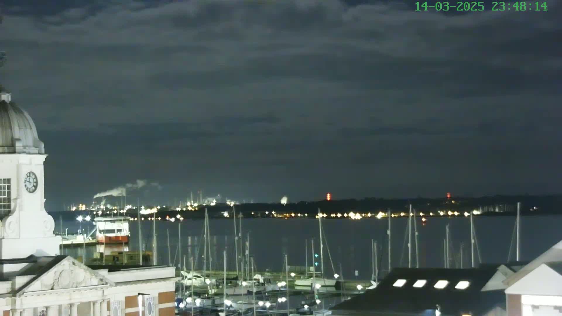 A nighttime view of a harbor filled with sailboats under a cloudy sky, with a clock tower and distant industrial lights visible.