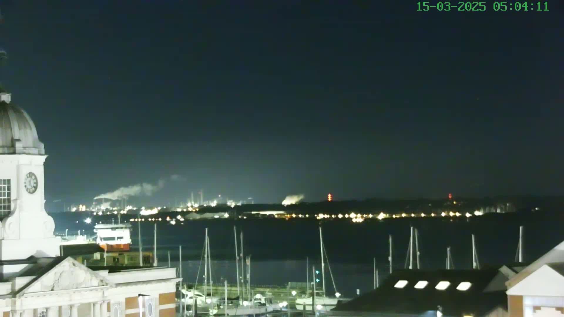 A nighttime view from a waterfront building shows a calm body of water with docked sailboats, an industrial area with smoke plumes in the distance, and a clear dark sky.