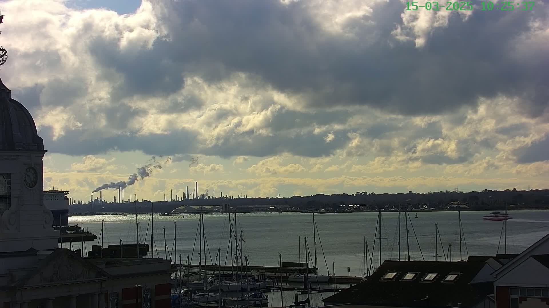 A partly cloudy sky overlooks a body of water filled with docked sailboats, with an industrial plant emitting smoke visible in the distance.