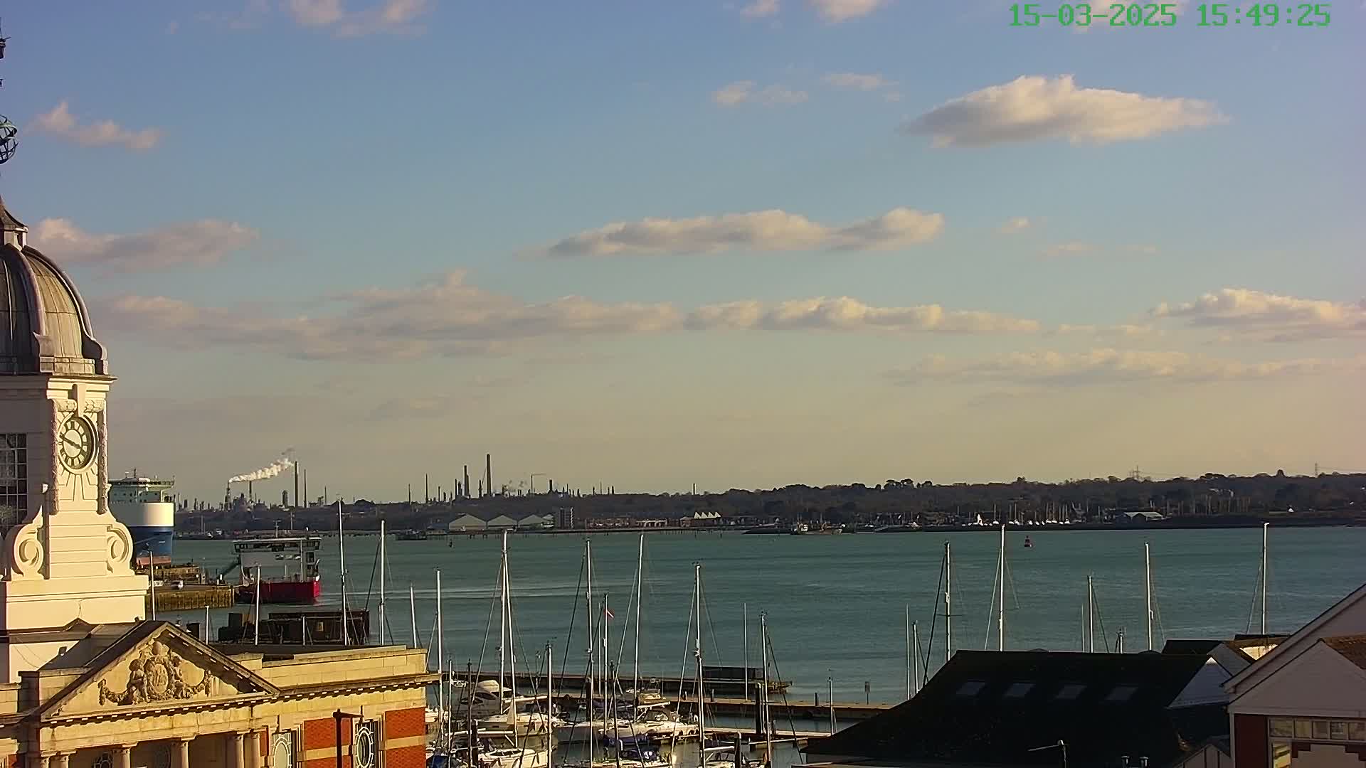 A clock tower overlooks a calm body of water with numerous sailboats docked, a distant industrial skyline under a partly cloudy sky.