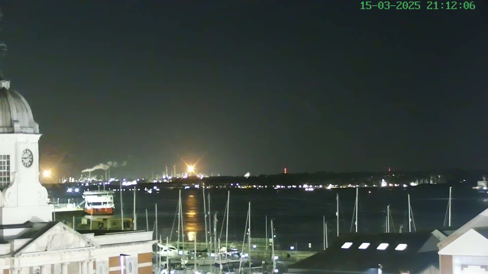 A nighttime view from atop a building shows a calm body of water with numerous masts of boats in a marina, an industrial area across the water, and a portion of a clock tower.