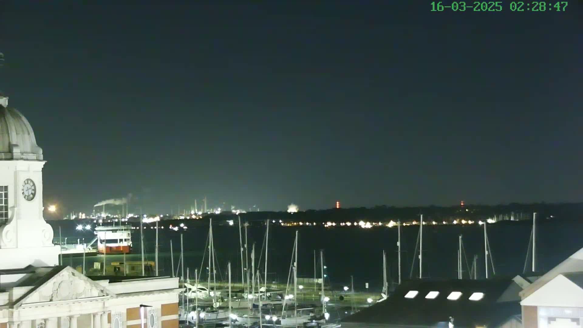 A nighttime view shows a harbor filled with sailboats, a clock tower, and an industrial area across the water under a clear, dark sky.