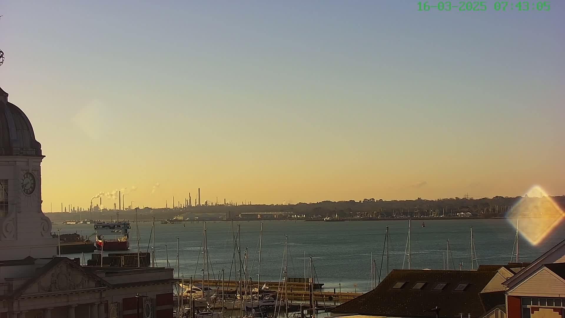 A calm body of water with numerous sailboats is seen from atop a building with a dome, under a clear, sunset sky; in the distance, an industrial area is visible on the horizon.