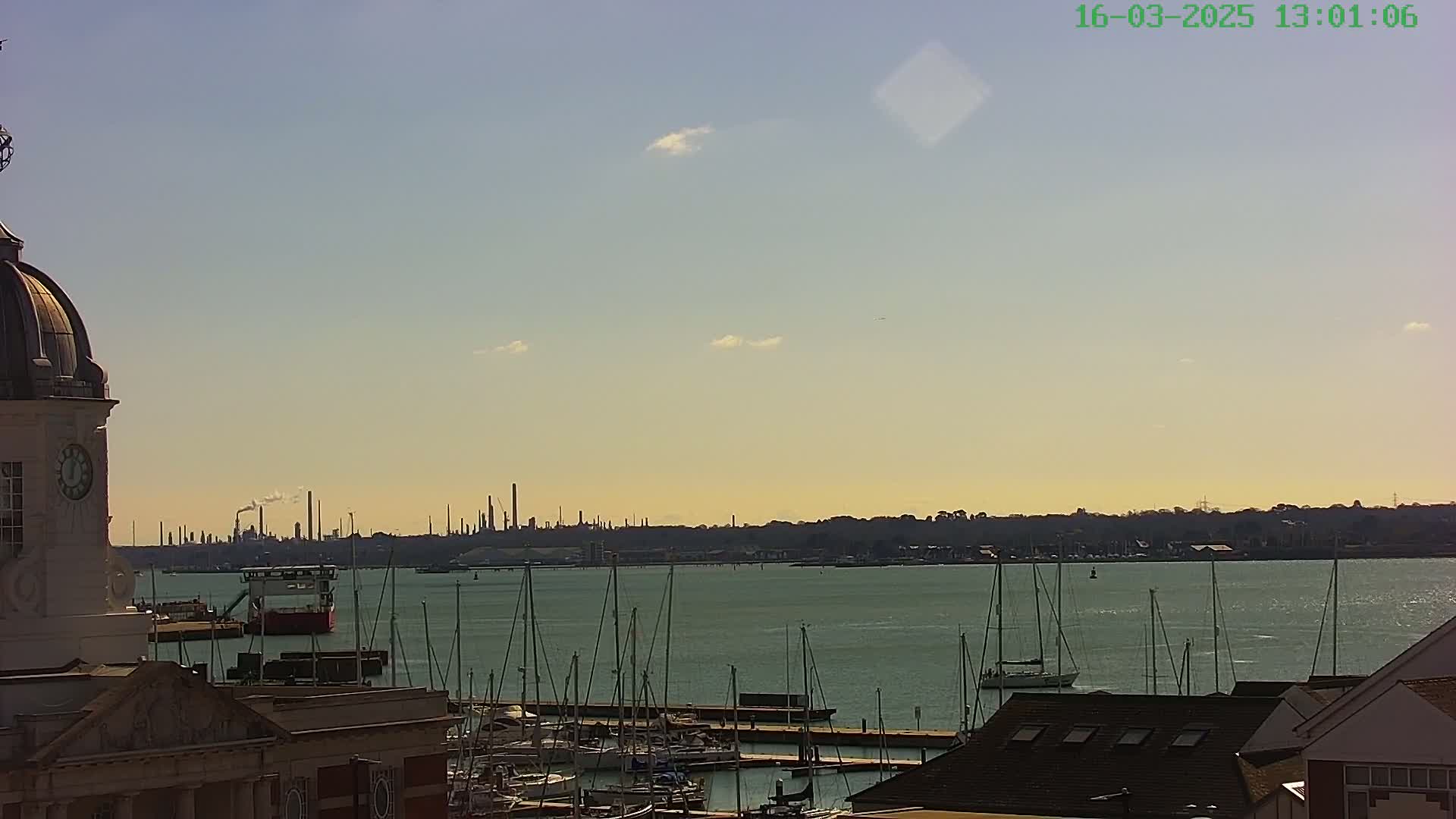 A view from a clock tower shows a calm body of water with numerous sailboats docked, a distant industrial skyline, and a mostly clear sky.