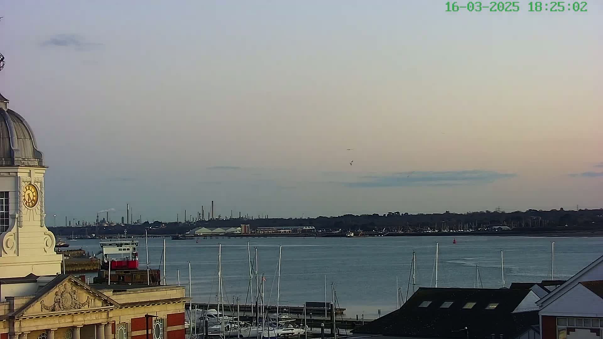 A clock tower overlooks a calm harbor with numerous sailboats docked, a distant industrial skyline under a pale, cloudy sky.