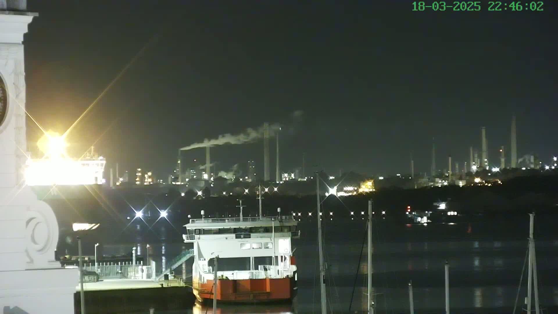 A red and white ferry is docked at night near a brightly lit industrial area emitting smoke, under a dark sky.