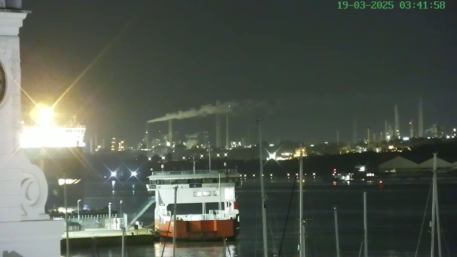 A docked ferry is illuminated at night, with a large industrial complex emitting smoke in the background, under a dark sky.