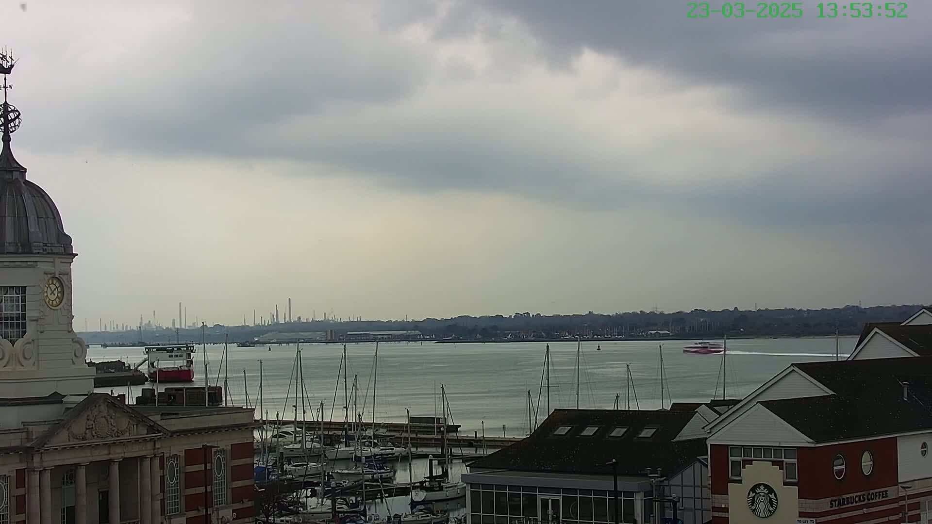 A harbor with many sailboats is seen from atop a building with a dome and clock under an overcast sky, with a distant industrial skyline visible across the water.